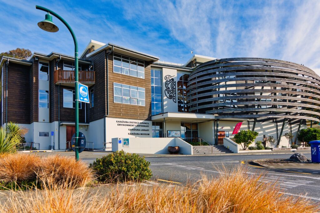 Modern curved building with dark timber and glass facade housing Kaikoura District Council, Environment Canterbury, Library and Museum. Green street lamp in foreground, native grasses, blue sky.